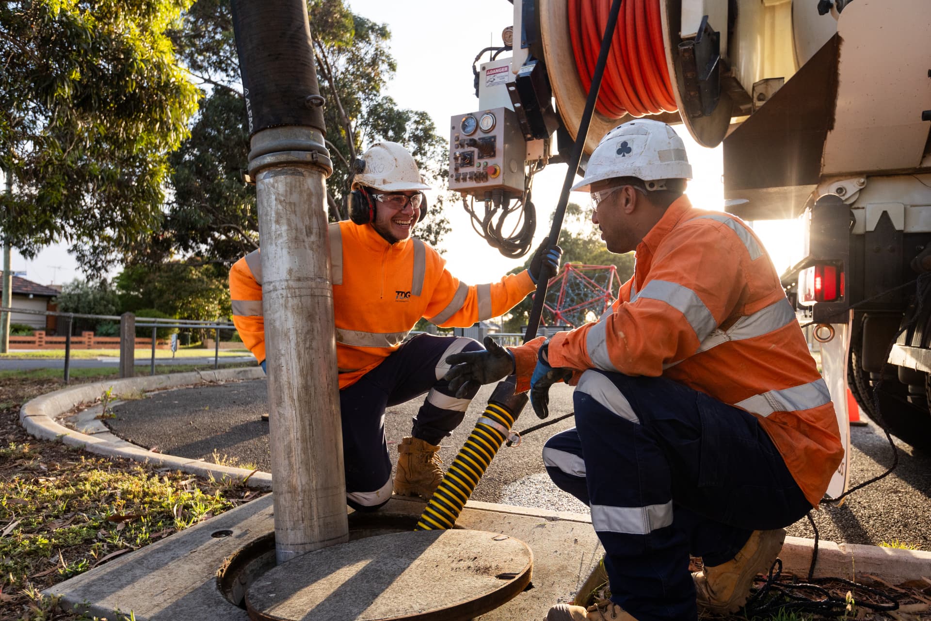 Two workers in hi-vis kneeling beside a manhole with drilling equipment at sunset